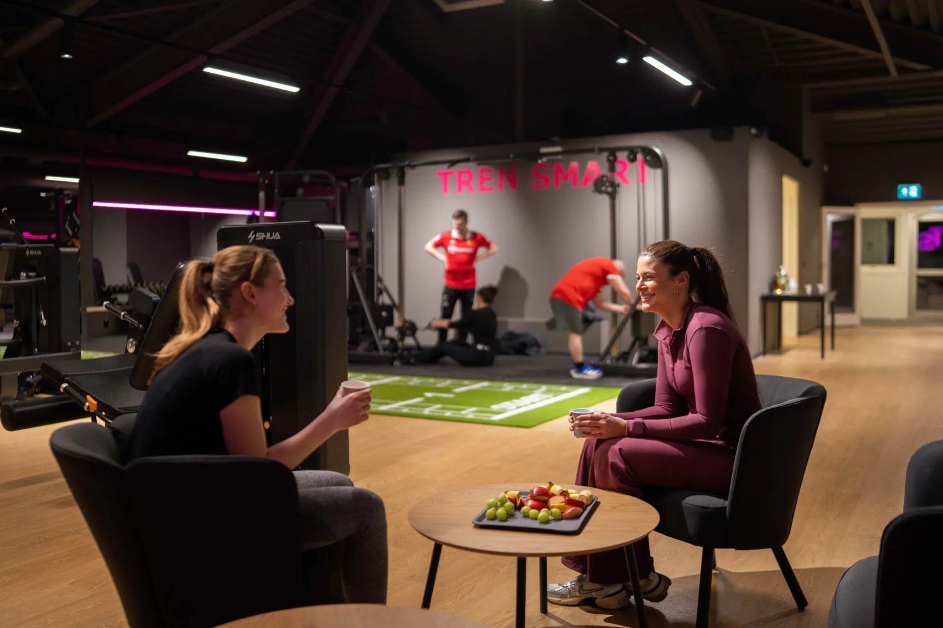 To medlemmer sitter i treningsstudioets lounge med frukt og kaffe, med TREN SMART-logoen i bakgrunnen